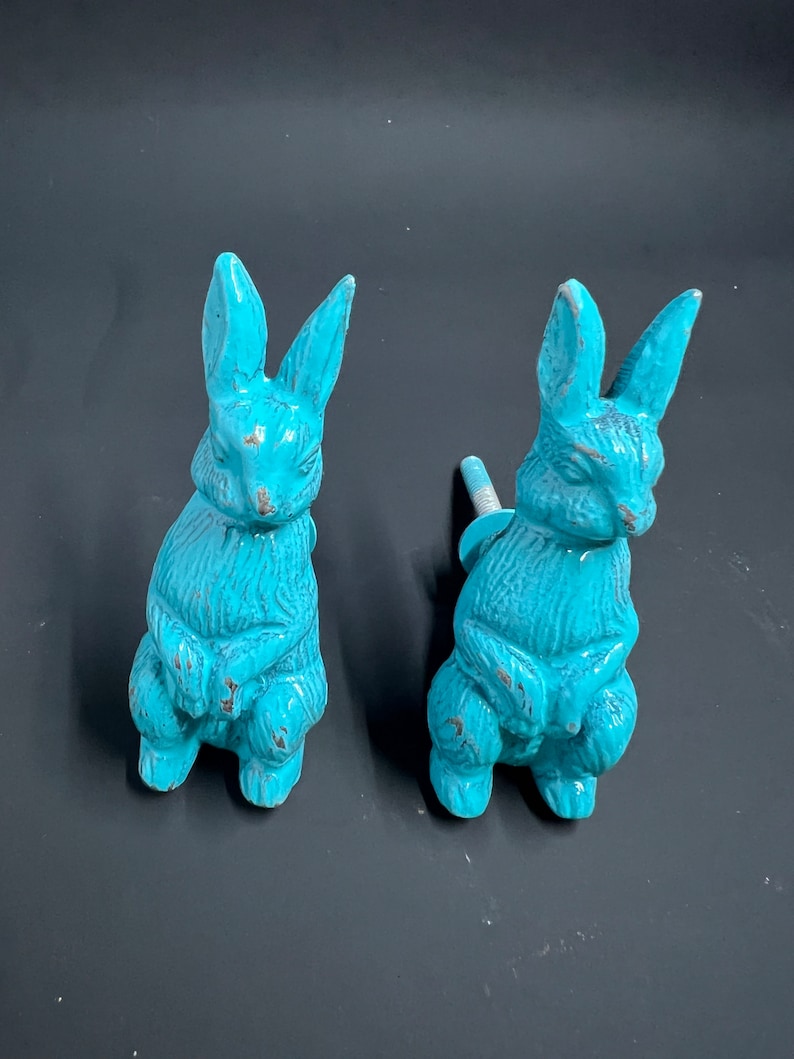 May include: Two turquoise blue ceramic rabbit figurines. The rabbits are sitting upright with their paws crossed. They have a distressed finish and are perfect for adding a touch of whimsy to your home decor.