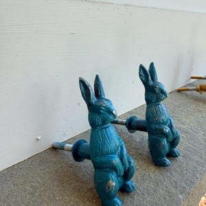 May include: Two teal-colored bunny-shaped cabinet pulls. The pulls are made of metal and have a distressed finish with gold accents. The bunnies are standing upright with their arms at their sides. The pulls are attached to a metal rod with a screw.