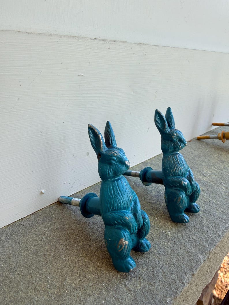 May include: Two teal-colored bunny-shaped cabinet pulls. The pulls are made of metal and have a distressed finish with gold accents. The bunnies are standing upright with their arms at their sides. The pulls are attached to a metal rod with a screw.