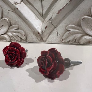 May include: Two red ceramic rose drawer pulls with silver screws. The rose pulls are in focus and the background is out of focus.