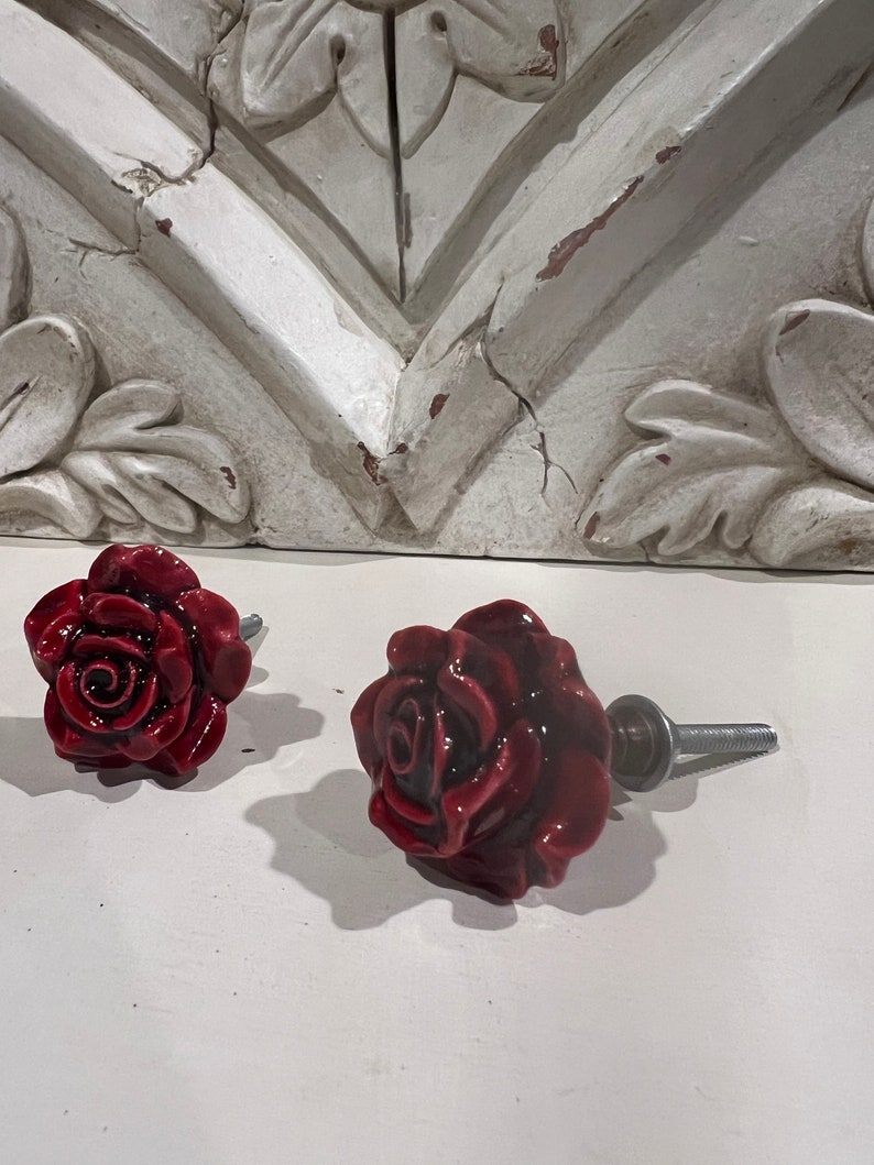 May include: Two red ceramic rose drawer pulls with silver screws. The rose pulls are in focus and the background is out of focus.
