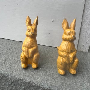 May include: Two yellow ceramic rabbit figurines. The rabbits are sitting upright with their ears perked up. They have a distressed finish and are a vintage style.
