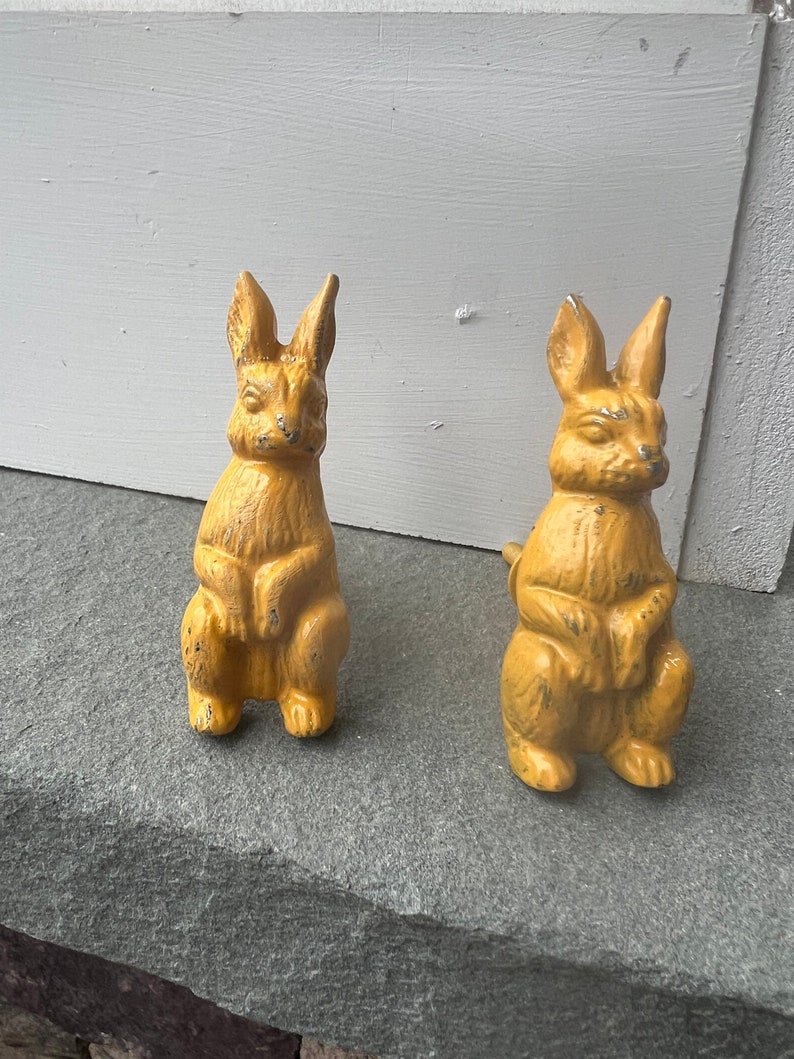 May include: Two yellow ceramic rabbit figurines. The rabbits are sitting upright with their ears perked up. They have a distressed finish and are a vintage style.
