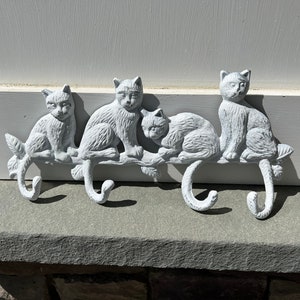 May include: White metal cat-shaped hooks on a branch. The hooks are designed to look like four cats sitting on a branch. The cats are all facing forward and have their tails curled around the branch. The hooks are a great way to add a touch of whimsy to any home.