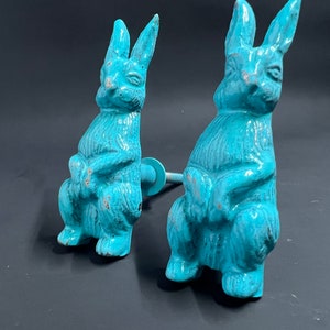 May include: Two turquoise blue ceramic rabbit figurines with a distressed finish. The rabbits are sitting upright with their paws crossed and have long ears. Each rabbit has a metal rod attached to the back for hanging.