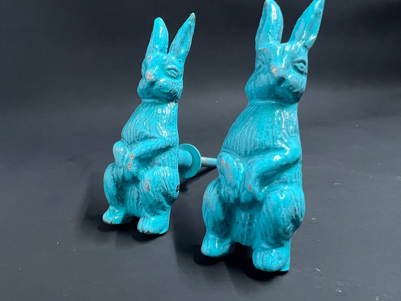May include: Two turquoise blue ceramic rabbit figurines with a distressed finish. The rabbits are sitting upright with their paws crossed and have long ears. Each rabbit has a metal rod attached to the back for hanging.
