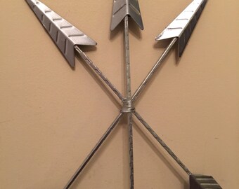 May include: A silver metal wall decor featuring four arrows arranged in a starburst pattern. The arrows are pointed and have a textured surface.