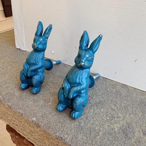 May include: Two teal-colored bunny-shaped cabinet knobs. The knobs are made of metal and have a distressed finish, giving them a vintage look. The bunnies are in a seated position with their ears upright. The knobs are mounted on a gray stone surface.