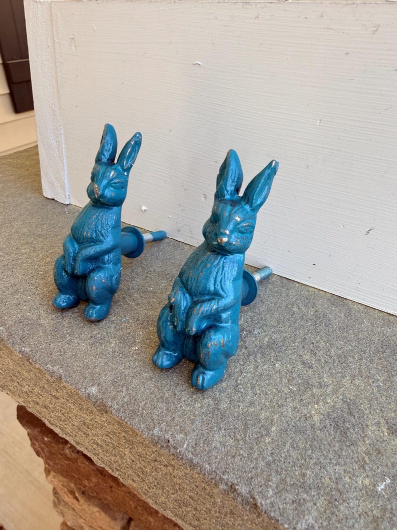 May include: Two teal-colored bunny-shaped cabinet knobs. The knobs are made of metal and have a distressed finish, giving them a vintage look. The bunnies are in a seated position with their ears upright. The knobs are mounted on a gray stone surface.