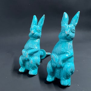 May include: Two turquoise blue ceramic bunny rabbit figurines. The bunnies are sitting with their paws crossed and have a distressed finish.