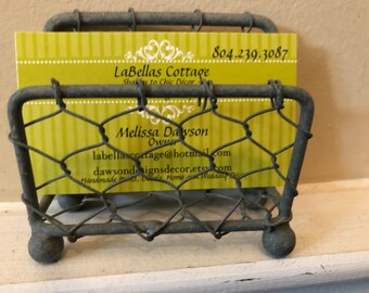 May include: A metal wire business card holder with a yellow business card inside. The card reads "LaBellas Cottage, Shabby to Chic Decor, 804.239.3087, Melissa Dawson, Owner, labellascottage@hotmail.com, dawsondesignsdecor.etsy.com, Handmade Items, Dolls, Home & Wedding Decor."