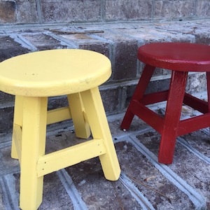 May include: Two wooden stools, one painted yellow and the other red, are sitting on a brick surface.