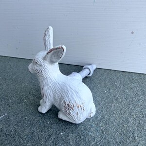 May include: A white rabbit-shaped cabinet knob with a distressed finish showing brown undertones. The rabbit is depicted in a seated position with upright ears and textured fur details. A metal bolt and nut extend from the back of the rabbit figure.
