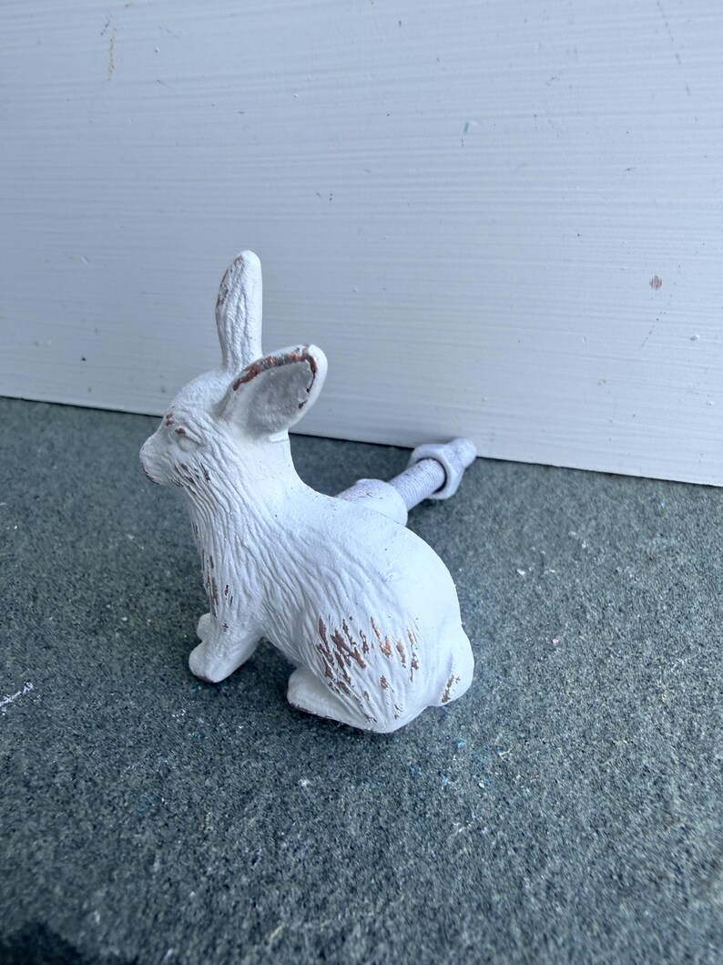 May include: A white rabbit-shaped cabinet knob with a distressed finish showing brown undertones. The rabbit is depicted in a seated position with upright ears and textured fur details. A metal bolt and nut extend from the back of the rabbit figure.