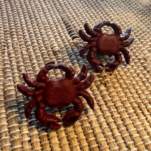 Op de afbeelding: Twee donkerrode krabfiguren met gedetailleerde klauwen en lichaamskenmerken. De krabben zijn geplaatst op een geweven, beige mat. De figuren zijn waarschijnlijk decoratieve items voor een nautisch thema.
