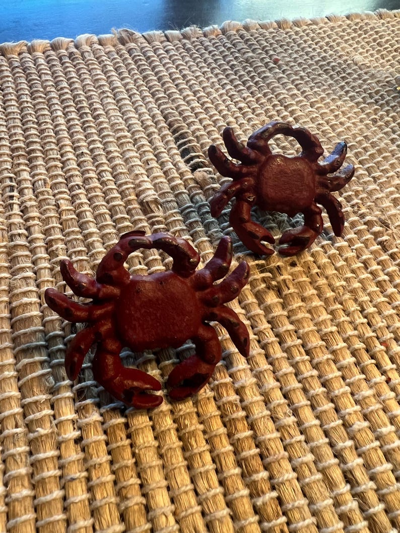 Op de afbeelding: Twee donkerrode krabfiguren met gedetailleerde klauwen en lichaamskenmerken. De krabben zijn geplaatst op een geweven, beige mat. De figuren zijn waarschijnlijk decoratieve items voor een nautisch thema.