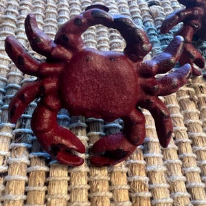May include: Close-up of a burgundy crab-shaped decorative object. The crab has multiple legs and claws, with black accents on the tips. The object is made of a textured material and is set against a woven background.