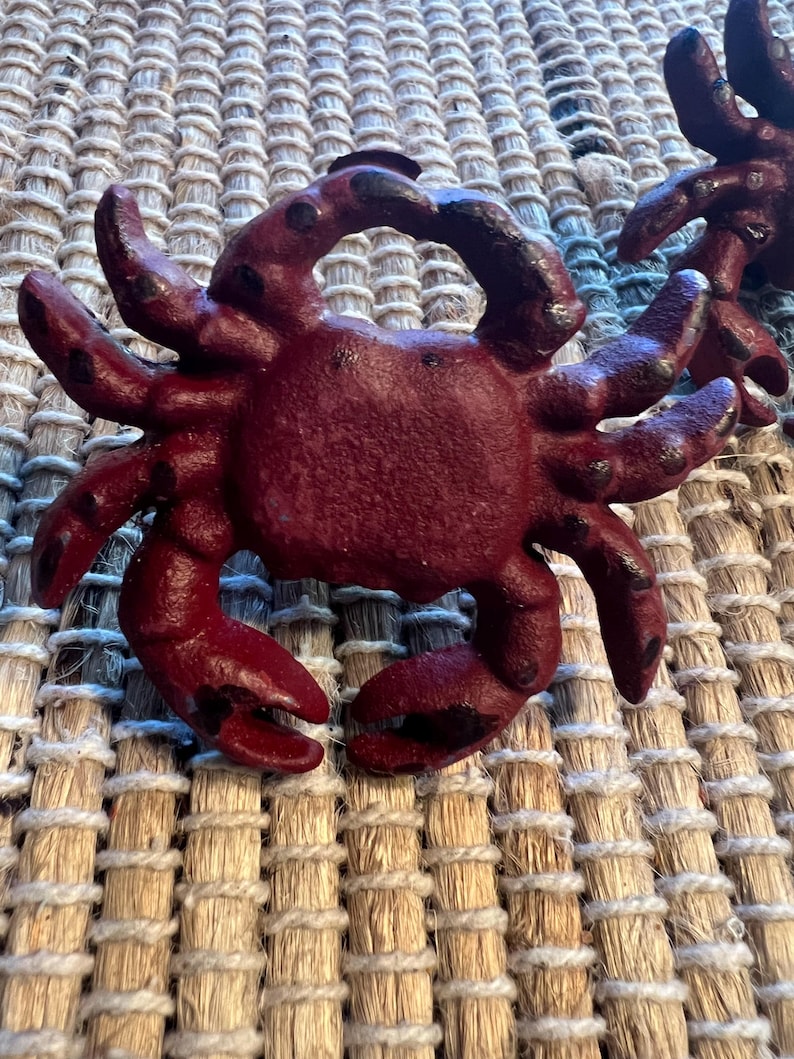 Op de afbeelding: Close-up van een bordeauxrode, krabvormige decoratie. De krab heeft meerdere poten en klauwen, met zwarte accenten op de uiteinden. Het object is gemaakt van een textuurrijk materiaal en staat tegen een geweven achtergrond.