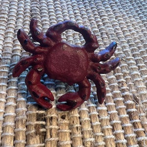 Op de afbeelding: Een bordeauxrode krabfiguur met gedetailleerde scharen en poten. De krab is gemaakt van een stevig materiaal en heeft een textuur. De figuur is een decoratief item, mogelijk voor een kust- of nautisch thema.