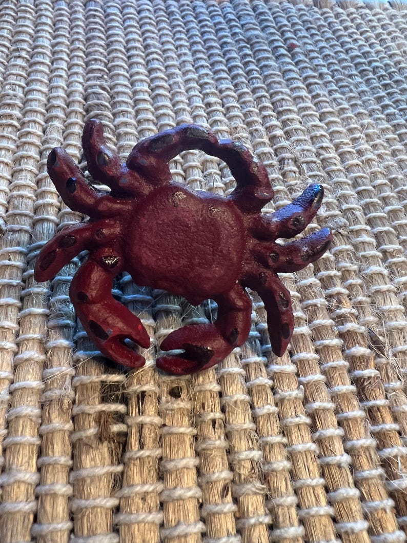 Op de afbeelding: Een bordeauxrode krabfiguur met gedetailleerde scharen en poten. De krab is gemaakt van een stevig materiaal en heeft een textuur. De figuur is een decoratief item, mogelijk voor een kust- of nautisch thema.