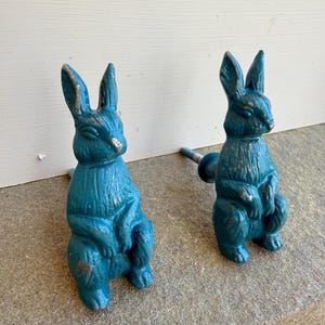 May include: Two teal-colored bunny-shaped cabinet knobs with a distressed finish. The knobs are cast in a sitting pose with their ears upright. The hardware is mounted on a stone surface, suggesting outdoor or garden use.