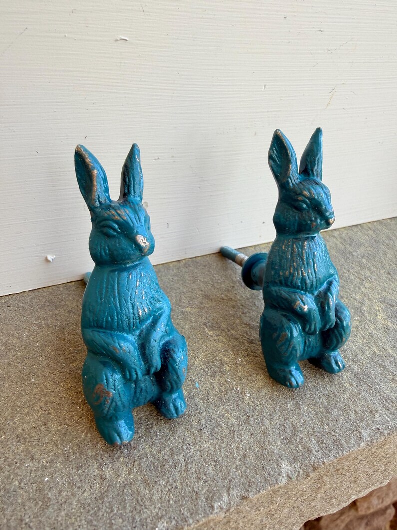 May include: Two teal-colored bunny-shaped cabinet knobs with a distressed finish. The knobs are cast in a sitting pose with their ears upright. The hardware is mounted on a stone surface, suggesting outdoor or garden use.