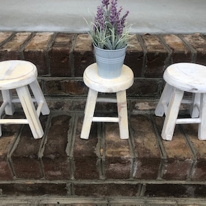 May include: Three white wooden stools with a distressed finish. The stools are arranged in a row on a brick surface. A small potted plant with purple flowers sits on the middle stool.
