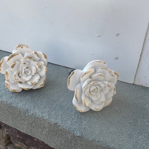 May include: Two white ceramic rose-shaped drawer pulls with gold accents. The pulls are on a gray stone surface.