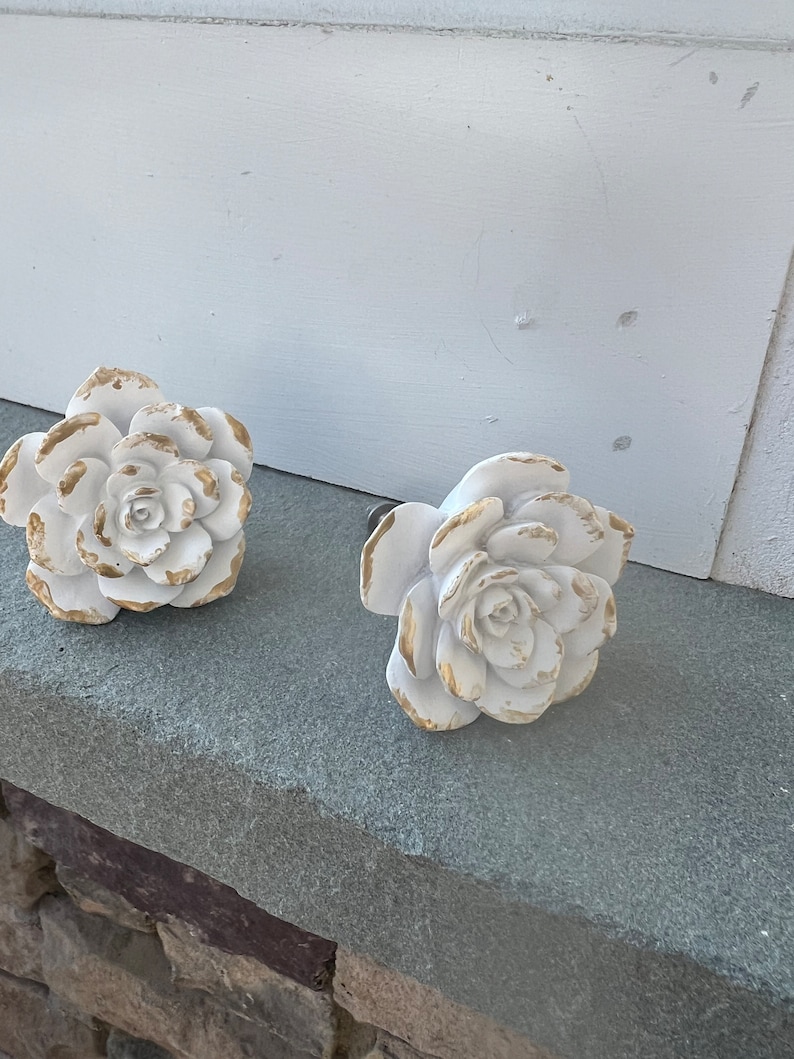 May include: Two white ceramic rose-shaped drawer pulls with gold accents. The pulls are on a grey stone surface.