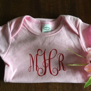 May include: Pink baby onesie with a red monogram "MHR" on the front. The onesie is folded and has a tag that says "Creations of Grace".