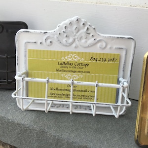 May include: White metal business card holder with a distressed finish and ornate design. The holder has a wire rack to hold business cards. The business card inside the holder reads "LaBellas Cottage, Shabby to Chic D&eacute;cor, labellascottage.etsy.com, 804.239.3087, Owner, labellascottag@hotmail.com, dawsondesigndecor.etsy.com, Handmade Items, Home D&eacute;cor and Wedding D&eacute;cor."