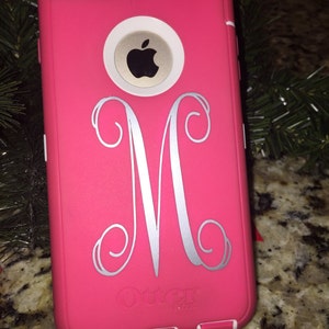 May include: A pink iPhone case with a white monogram "M" and the OtterBox logo. The case has a black camera lens and a white Apple logo.