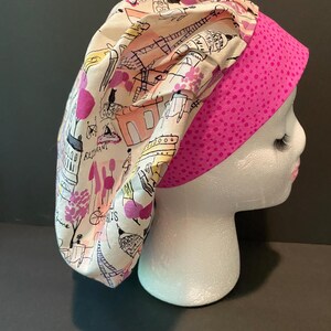 Può includere: Un cappello da lavoro bianco e rosa con una stampa a tema parigino. Il cappello presenta un bordo a pois rosa e uno sfondo bianco con illustrazioni nere e rosa di edifici, biciclette e altri monumenti parigini.