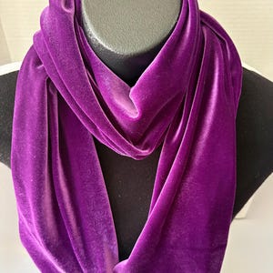 May include: A vibrant purple velvet infinity scarf draped over a black mannequin head. The scarf's soft texture and rich color are highlighted, showcasing its elegant design. This accessory is perfect for adding a touch of luxury to any outfit.