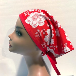 Puede incluir: Un pañuelo rojo con un estampado floral blanco. El pañuelo se ata en la parte posterior con cintas rojas. La tela tiene un diseño floral blanco. El pañuelo se muestra en una cabeza de maniquí.