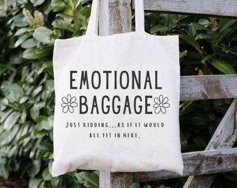 May include: A cream-colored tote bag with black text that reads "EMOTIONAL BAGGAGE" with a flower graphic. Below the main text is the phrase "JUST KIDDING...AS IF IT WOULD ALL FIT IN HERE."
