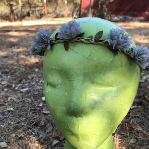 May include: A purple and green flower crown with a woven base. The crown is made of artificial flowers and is worn on a green mannequin head.