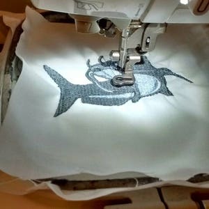 May include: An embroidery machine in operation, stitching a cartoon fish design onto white fabric. The fish is primarily gray with blue accents. The machine's needle is positioned over the fabric, creating the embroidered image.