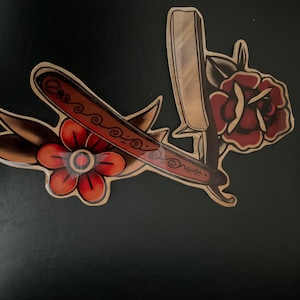 May include: A vintage-style tattoo design featuring a straight razor and two red flowers with green leaves. The razor is brown with a gold-colored handle and a decorative pattern.