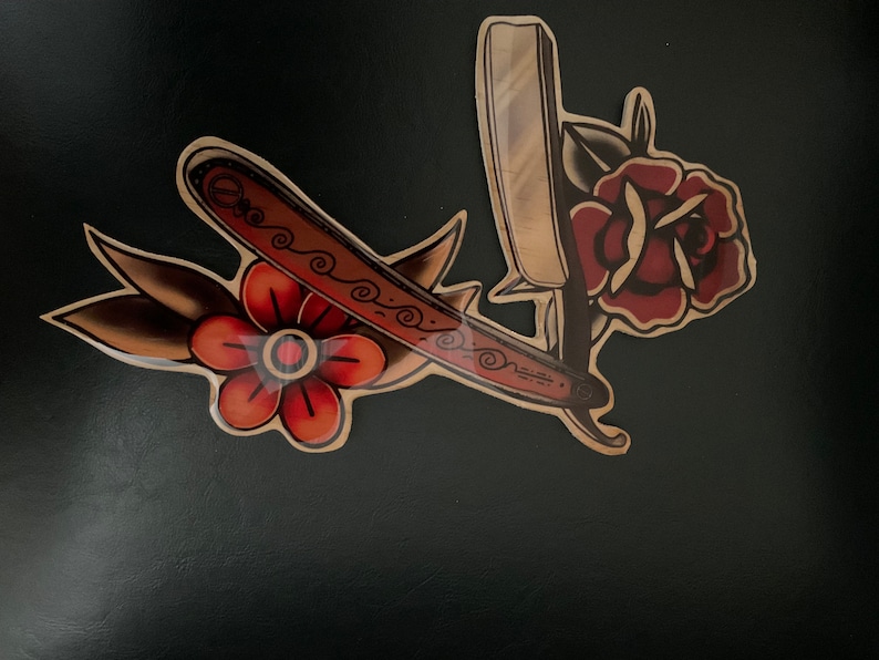 May include: A vintage-style tattoo design featuring a straight razor and two red flowers with green leaves. The razor is brown with a gold-colored handle and a decorative pattern.