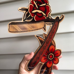 May include: A wooden wall art piece featuring a traditional tattoo style design of a straight razor and two red roses. The razor is detailed with a black and white pattern.