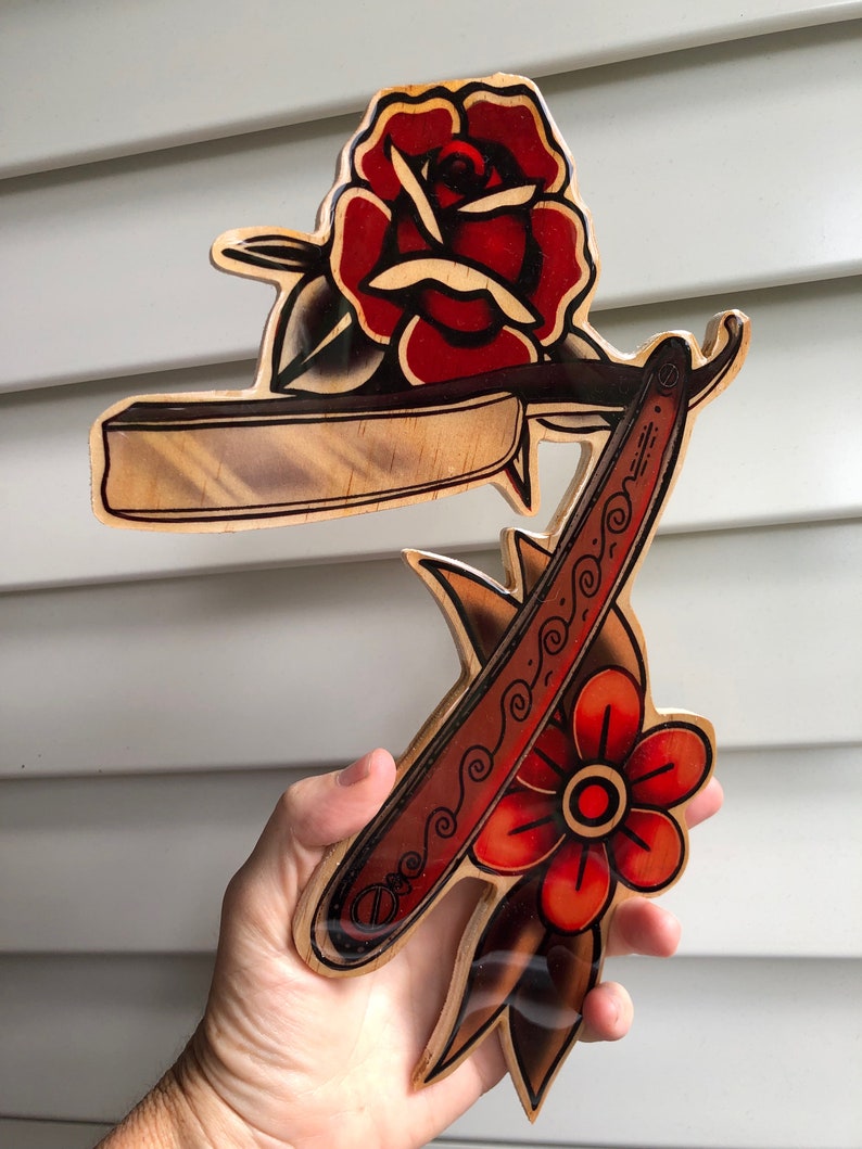 May include: A wooden wall art piece featuring a traditional tattoo style design of a straight razor and two red roses. The razor is detailed with a black and white pattern.