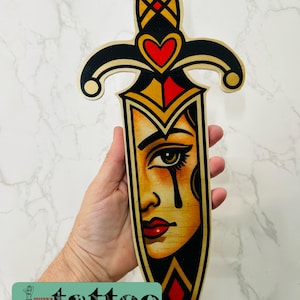 Puede incluir: Arte de pared de madera con forma de daga que presenta una ilustración estilizada del rostro de una mujer. La daga tiene un mango negro y dorado con detalles de corazón y diamante. El texto "cheeky tattoo" está en la parte inferior.