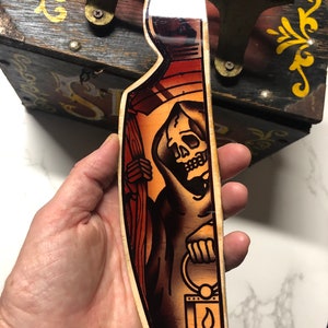 May include: A wooden plaque with a hand-painted illustration of a skeletal figure in a hooded robe holding a lantern. The figure is depicted in a traditional tattoo style with black outlines and vibrant colors. The background is a dark brown wood.