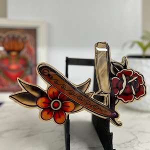 May include: A wooden wall decor featuring a vintage style tattoo design of a straight razor and a rose with a red, yellow, and green color scheme.