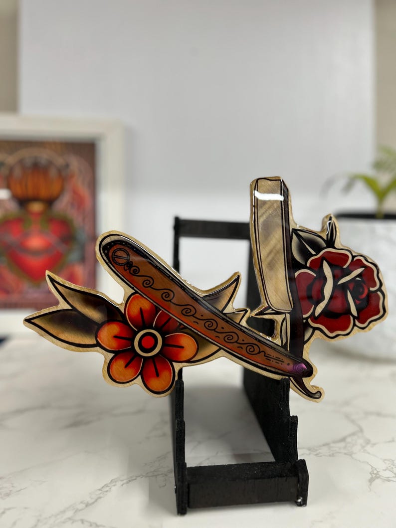 May include: A wooden wall decor featuring a vintage style tattoo design of a straight razor and a rose with a red, yellow, and green color scheme.