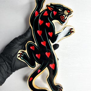 Op de afbeelding: Houten uitgesneden zwarte panter met rode hartaccenten. De panter heeft een felle uitdrukking, witte klauwen en een gouden omtrek. De uitgesneden figuur wordt vastgehouden door een gehandschoende hand.