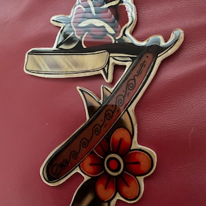 May include: A vintage-style tattoo design featuring a straight razor with a decorative handle and a rose with a single flower below. The design is printed on a sticker.
