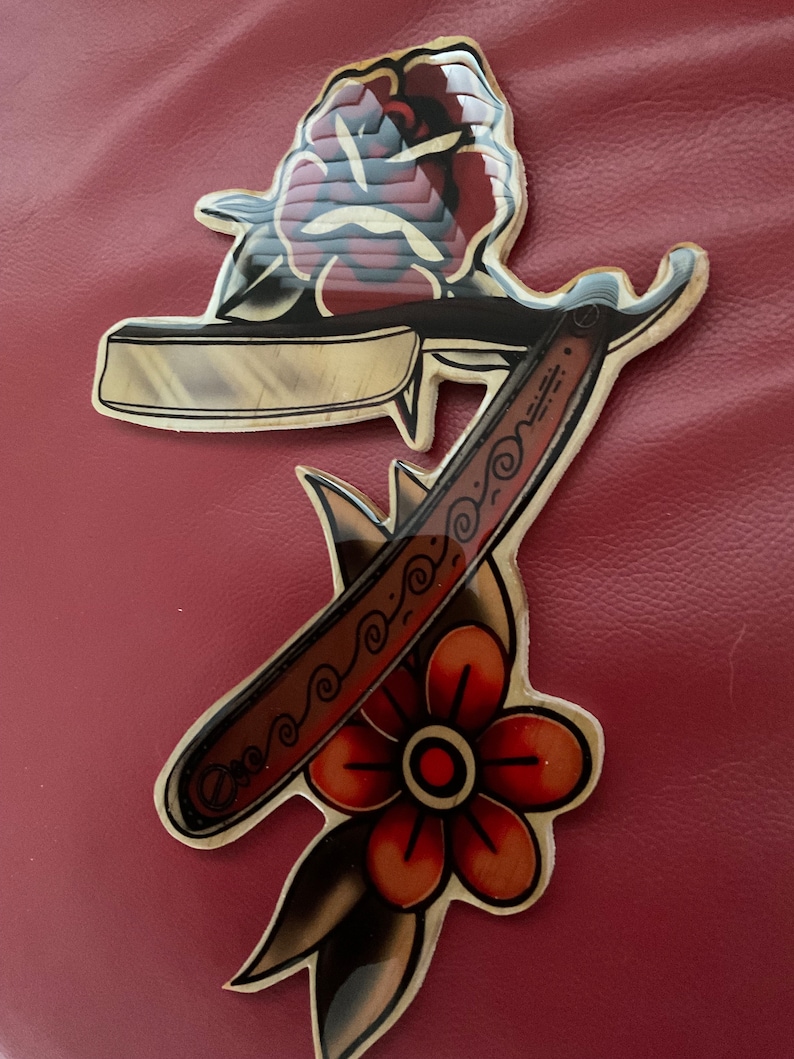 May include: A vintage-style tattoo design featuring a straight razor with a decorative handle and a rose with a single flower below. The design is printed on a sticker.