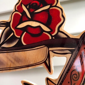 May include: A wooden wall art piece with a red rose and an anchor. The rose is painted in a traditional tattoo style with black outlines and red and white shading. The anchor is painted in a similar style with brown, black, and white shading.